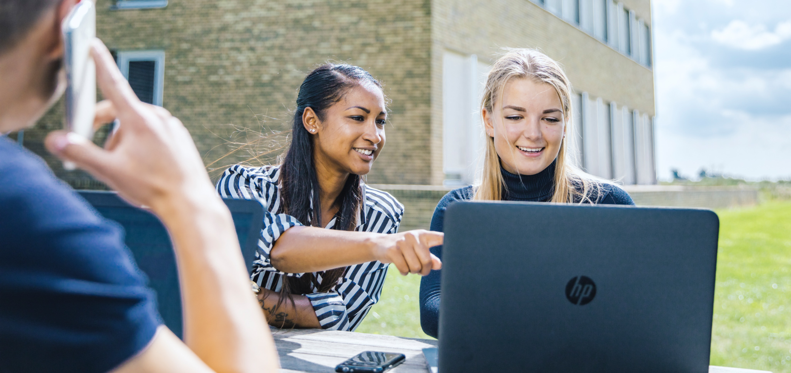 twee vrouwen zitten lachend achter hun laptop aan een picknicktafel achter het kantoorgebouw van hamilton bright