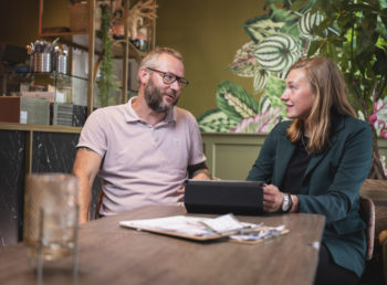 mannelijke horecamanager in roze polo in gesprek met een vrouwelijke junior rayonmanager in groen colbert in een restaurant