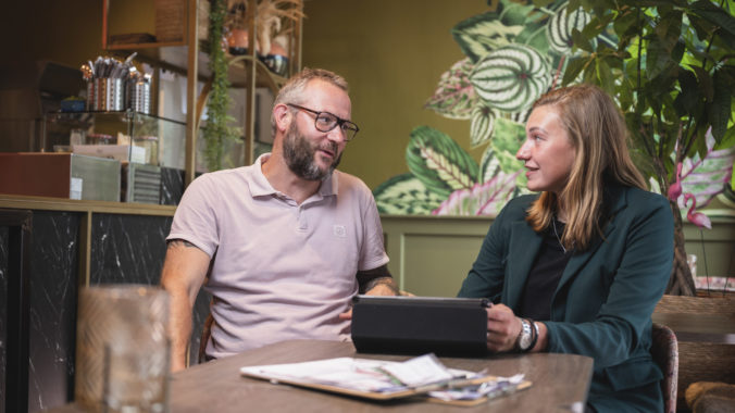 mannelijke horecamanager in roze polo in gesprek met een vrouwelijke junior rayonmanager in groen colbert in een restaurant