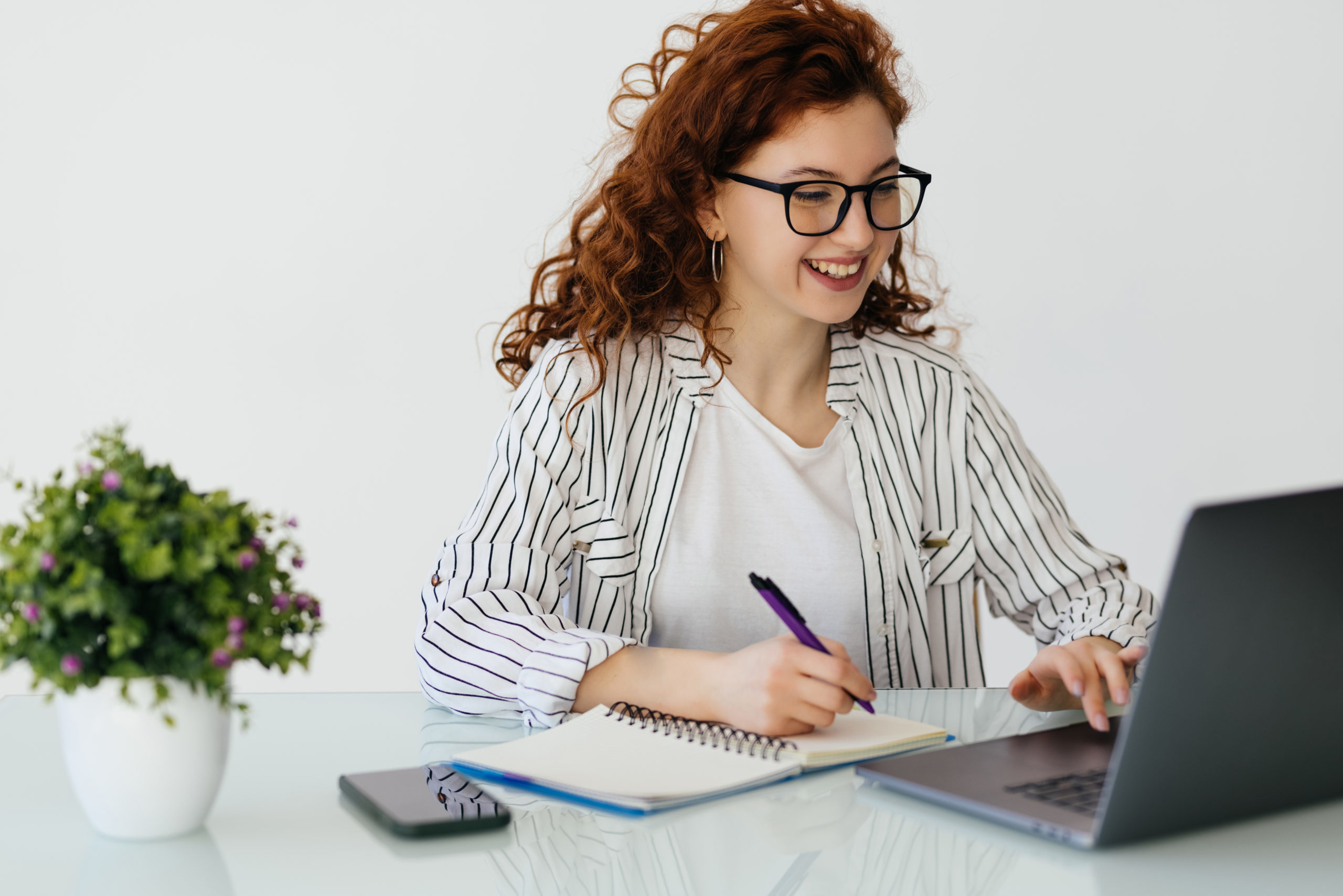 roodharige vrouw met bril analyseert documenten met een rekenmachine op een laptop aan een wit bureau